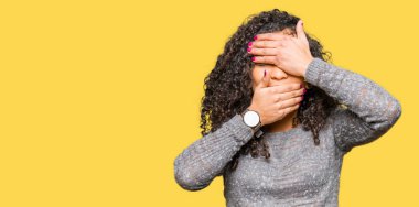 Kıvırcık saçlı genç güzel kadın gri kazak giyen gözleri ve ağzı elleriyle kapamış, şaşırmış ve şok etmiş. Duyguyu gizleme