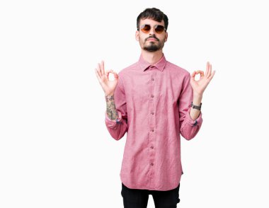 Genç yakışıklı adam giyen güneş gözlüğü izole arka plan üzerinde dinlenmek ve gülümseyen gözlerle parmak ile meditasyon jest yapıyor kapalı. Yoga kavramı.