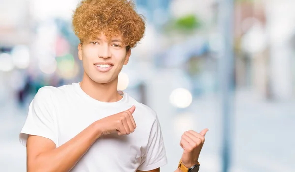 カジュアルな白いTシャツを着たアフロヘアの若いハンサムな男は 手と親指で後ろを指し 自信を持って微笑む — ストック写真