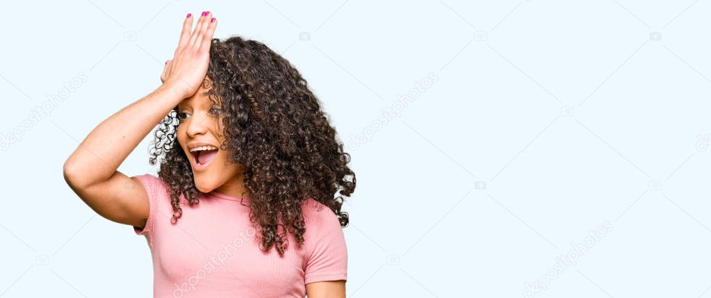 Young beautiful woman with curly hair wearing pink t-shirt surprised with hand on head for mistake, remember error. Forgot, bad memory concept.