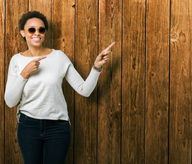 Güzel genç Afro-Amerikan kadın gülümseyerek ve iki elleri ve parmakları ile tarafına işaret kamera bakarak izole arka plan üzerinde güneş gözlüğü takıyor.