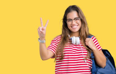 Mutlu yüz zafer işareti yaparken kameraya göz kırpıyor ile gülümseyen izole arka plan üzerinde kulaklık ve sırt çantası giyen genç güzel esmer öğrenci kadın. İki numaralı.