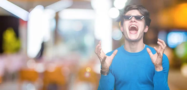 Young man wearing funny thug life glasses over isolated background crazy and mad shouting and yelling with aggressive expression and arms raised. Frustration concept.