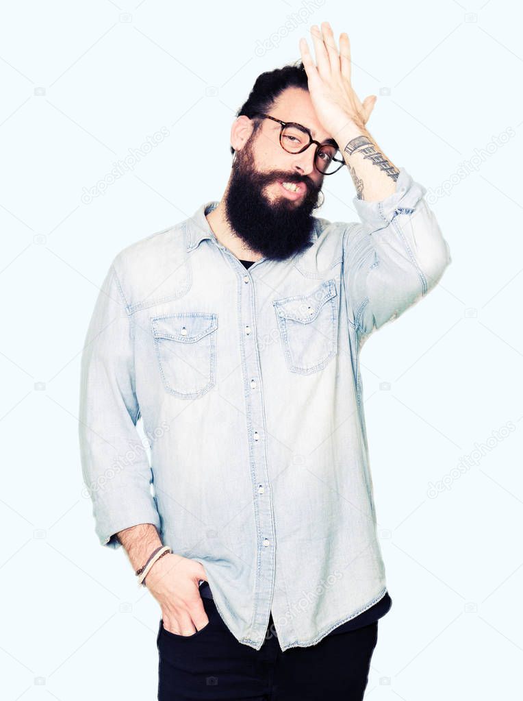 Young hipster man with long hair and beard wearing glasses surprised with hand on head for mistake, remember error. Forgot, bad memory concept.