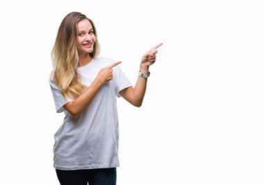 Gülümseyen ve iki elleri ve parmakları ile tarafına işaret kamera bakarak izole arka plan üzerinde rahat beyaz t-shirt giyen genç güzel sarışın kadın.