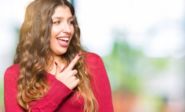 Kırmızı kazak giyen genç güzel kadın yüzünde mutlu ve doğal ifade ile yan yana el ve parmak ile işaret yüz bir gülümseme ile neşeli