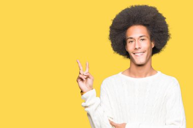 Afro saç ile genç Afrikalı Amerikan adam kış kazak gülümseyen mutlu yüz zafer işareti yapıyor kamera göz kırpıyor ile sırıtıyor. İki numara.