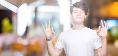 Casual beyaz t-shirt izole arka plan üzerine dinlenmek giydiği ve gözleri ile gülümseyen yakışıklı delikanlı parmak ile meditasyon jest yapıyor kapalı. Yoga kavramı.