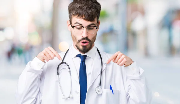 若い医者分離オーバー病院のコートを身に着けている男がダウン表示広告 驚いた顔 口を開けて指でポインティングを背景します — ストック写真