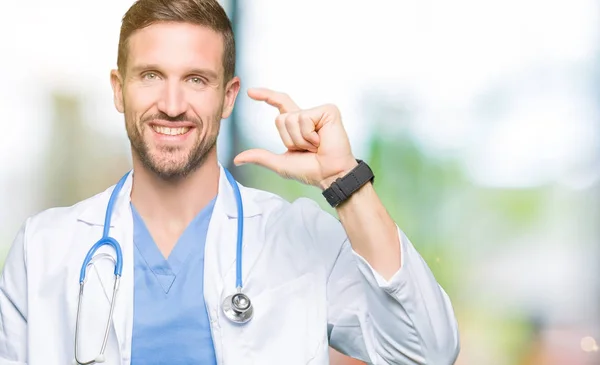 英俊的医生穿医疗制服在孤立的背景微笑和自信的手势与手做大小标志用手指 而看和相机 测量概念 — 图库照片
