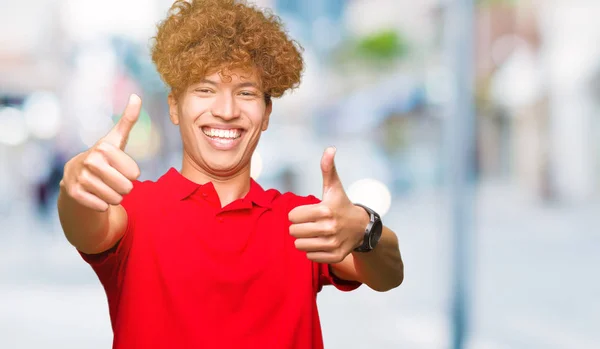 赤いTシャツを着たアフロヘアの若いハンサムな男は 手で肯定的なジェスチャーを行うことを承認し 親指は笑顔で成功のために幸せです カメラを見て 勝者のジェスチャー — ストック写真
