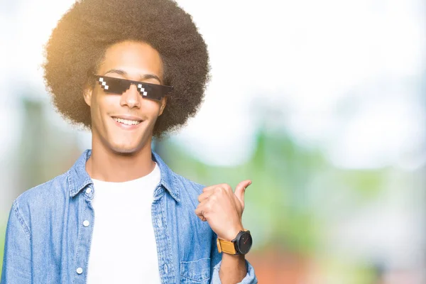 Young african american man with afro hair wearing thug life glasses smiling with happy face looking and pointing to the side with thumb up.