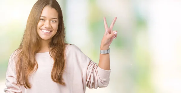 Young beautiful brunette woman wearing pink sweatshirt over isolated background smiling with happy face winking at the camera doing victory sign. Number two.