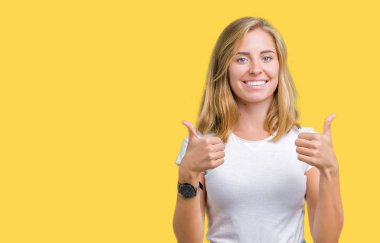 El ile pozitif jest yapıyor izole arka plan başarı işareti üzerinde rahat beyaz t-shirt giyen güzel ve genç birbayan başparmak gülümseyen ve mutlu. Neşeli bir ifadeyle, kazanan jest kameraya bakıyor.