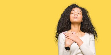 Kıvırcık saçlı ile gülümseyen rahat kazak giyen genç güzel kız göğüs kapalı gözler ve yüzünde minnettar jest ile eller. Sağlık kavramı.