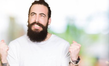 Uzun saç ve sakal gülümseyen ve başarı için çığlık casual beyaz t-shirt çok mutlu ve heyecanlı kaldırdı, silah ile kazanan jest yapıyor giyen genç hipster adam. Kutlama kavramı.