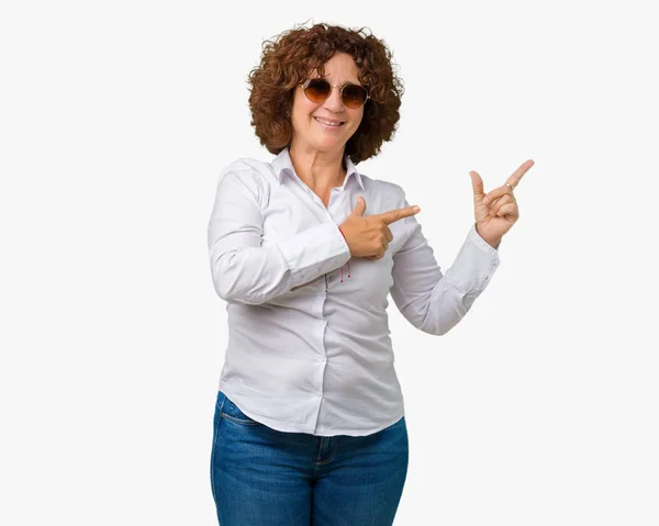 笑顔で側に つの手と指で指しているカメラを見て孤立の背景にサングラスをかけて美しい中間エイガー シニア ビジネス女性 — ストック写真