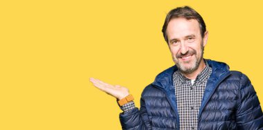 Orta yaş yakışıklı adam kış ceket giyen neşeli sunan gülümseyen ve el kamera bakarak avuç ile işaret.