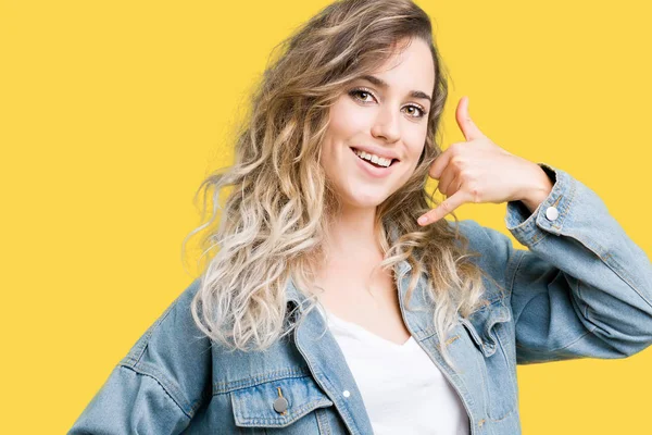 Beautiful young blonde woman wearing denim jacket over isolated background smiling doing phone gesture with hand and fingers like talking on the telephone. Communicating concepts.