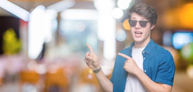 Yakışıklı delikanlı gülümseyen ve iki elleri ve parmakları ile tarafına işaret kamera bakarak izole arka plan üzerinde güneş gözlüğü takıyor.