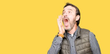 Orta yaş yakışıklı adam kış yelek giyen bağırıyor ve ağız üzerinde el ile yan gürültülü çığlık. İletişim konsepti.