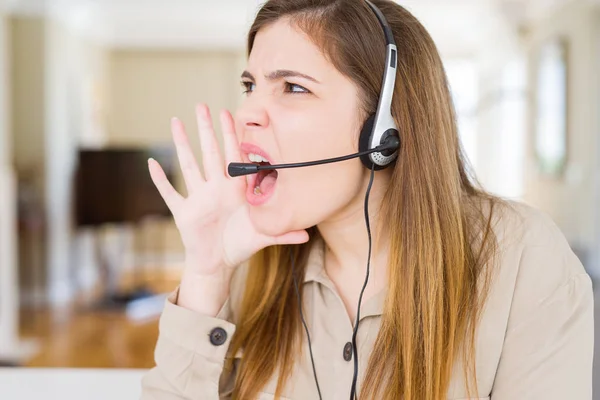 Beautiful young operator woman wearing headset at the office shouting and screaming loud to side with hand on mouth. Communication concept.