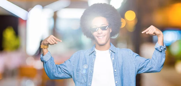 Young african american man with afro hair wearing thug life glasses looking confident with smile on face, pointing oneself with fingers proud and happy.