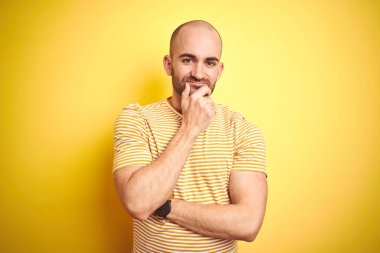 Sakallı genç kel adam sarı izole arka plan üzerinde rahat çizgili t-shirt giyen kamera çapraz kollar ve el çene üzerinde kaldırdı gülümseyerek emin görünümlü. Olumlu düşünme.
