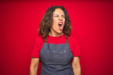 Orta yaş yaşlı kadın kırmızı izole arka plan üzerinde önlük üniforma giyen kızgın ve deli çığlık sinirli ve öfkeli, öfke ile bağırarak. Öfke ve agresif kavram.