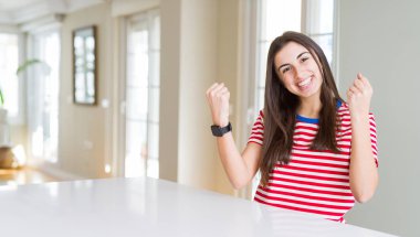 Rahat çizgili tişört giyen güzel genç kadın, kollarını kaldırıp açık gözlerle başarıiçin şaşırmış ve şaşırmış. Kazanan kavramı.