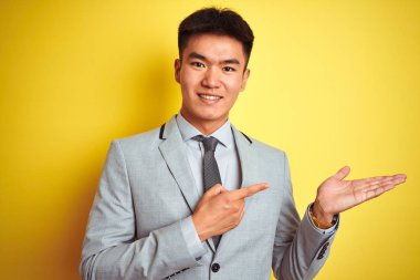 Takım elbise giyen ve kravat takan Asyalı çinli işadamı izole sarı arka plan üzerinde duran şaşırttı ve el ile sunarken kameraya gülümseyerek ve parmak ile işaret.