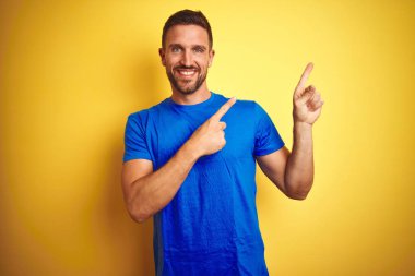 Genç yakışıklı adam sarı izole arka plan üzerinde rahat mavi t-shirt giyen gülümseyerek ve yan iki el ve parmakları ile işaret kamera bakarak.