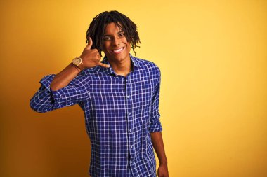 Yalnız sarı arka plan üzerinde duran rahat gömlek giyen dreadlocks ile Afro adam telefonda konuşurken gibi el ve parmakları ile telefon jest yapıyor gülümseyerek. İletişim kavramları.