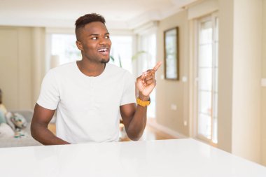 Yakışıklı afro-amerikan adam evde yüzünde büyük bir gülümseme ile beyaz masada, kameraya bakarak yan el ve parmak ile işaret.