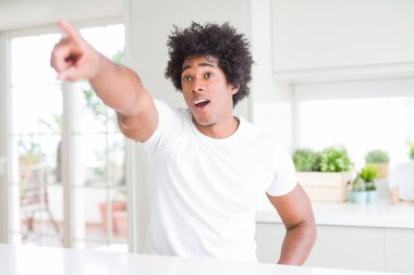 Genç afro-amerikan adam evde oturan rahat beyaz t-shirt giyen önünde sürpriz parmak ile işaret, açık ağız şaşkın ifade, ön bir şey