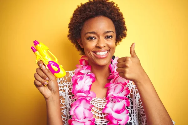Young african american woman with afro hair wearing flower hawaiian lei and water gun happy with big smile doing ok sign, thumb up with fingers, excellent sign