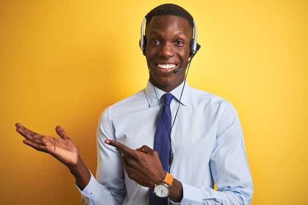 African american operator man working using headset over isolated yellow background amazed and smiling to the camera while presenting with hand and pointing with finger.