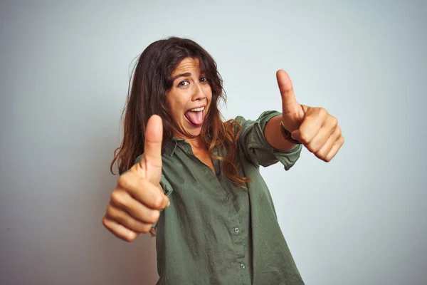 Young beautiful woman wearing green shirt standing over grey isolated background approving doing positive gesture with hand, thumbs up smiling and happy for success. Winner gesture.