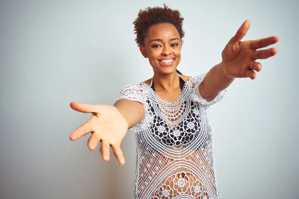 Young african american woman with afro hair wearing a bikini over white isolated background looking at the camera smiling with open arms for hug. Cheerful expression embracing happiness.