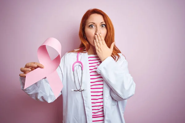 Young redhead doctor woman holding cancer ribbon over pink isolated background cover mouth with hand shocked with shame for mistake, expression of fear, scared in silence, secret concept