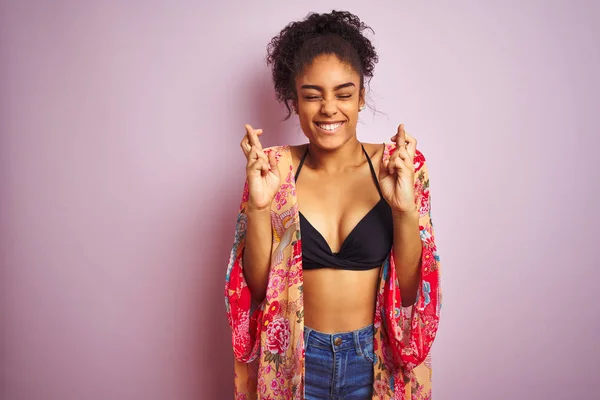 American woman on holiday wearing bikini and colorful dress over isolated pink background gesturing finger crossed smiling with hope and eyes closed. Luck and superstitious concept.