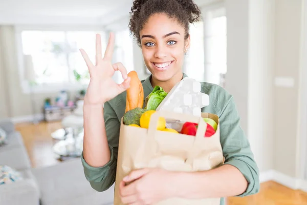 Young african american girl holding paper bag of groceries from supermarket doing ok sign with fingers, excellent symbol