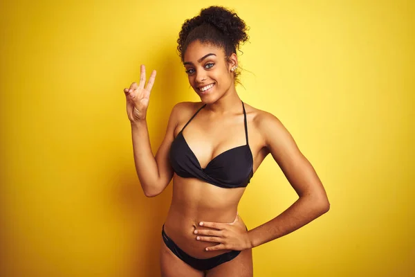 African american woman on vacation wearing bikini standing over isolated yellow background smiling looking to the camera showing fingers doing victory sign. Number two.