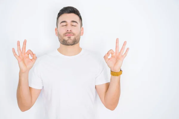 年轻的英俊男子穿着白色休闲白色T恤 在白色孤立的背景放松和微笑 闭上眼睛用手指做冥想手势 瑜伽概念 — 图库照片