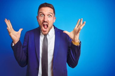 Mavi izole arka plan üzerinde genç yakışıklı iş adamı deli kutluyor ve kollarını kaldırdı ve açık gözleri heyecanlı çığlık ile başarı için hayran. Kazanan kavramı