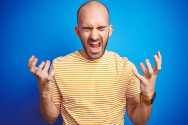 Sakallı genç kel adam mavi izole arka plan üzerinde rahat t-shirt giyen silah kaldırdı ve kapalı gözleri heyecanlı çığlık ile başarı için deli ve deli kutluyor. Kazanan kavramı