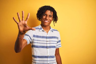 Afro amerikan adam dreadlocks izole sarı arka plan üzerinde çizgili gömlek giyen gösteren ve parmakları ile işaret ederken emin ve mutlu gülümseyerek beş.