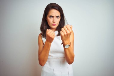 Beyaz izole arka plan üzerinde duran elbise giyen genç güzel kadın yumruk savunma jesti ile mücadele etmeye hazır, kızgın ve üzgün yüz, sorun korkuyor