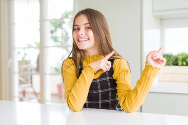 Genç güzel sarışın çocuk kız evde rahat sarı kazak giyen gülümseyerek ve yan iki el ve parmakları ile işaret kamera bakıyor.