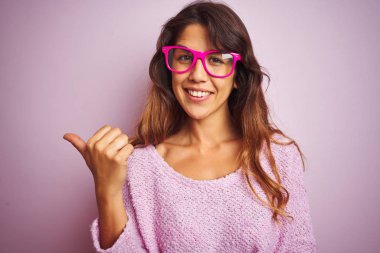 Pembe izole arka plan üzerinde duran moda gözlüğü takan genç güzel kadın mutlu yüz görünümlü ve başparmak ile yan işaret gülümseyerek.